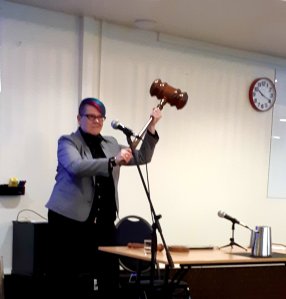 A person (Darusha) with pink and blue stands behind a desk and hair holds a large wooden mace aloft.